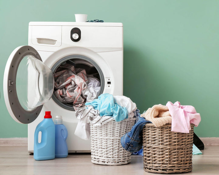 Geopende wasmachine met kleurrijke was, twee rieten wasmanden en wasmiddelflessen op de vloer